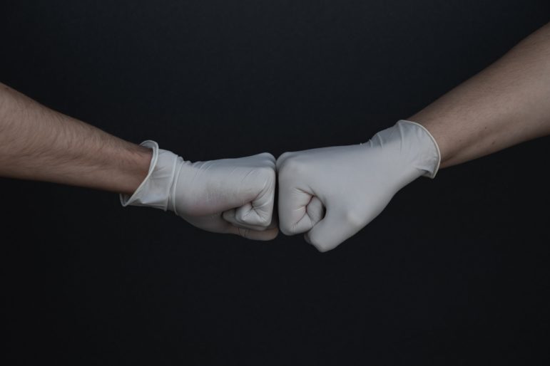 fist bump against dark background. Both fists have surgical latex gloves on.